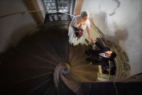 Braut läuft im Eberbacher Schloss die gewendelte Treppe hinauf. In einer Hand hat sie ihren Brautstrauß, mit der anderen hält sie zärtlich die Hans ihres Ehemannes der ihr folgt.