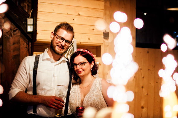Brautpaar steht vor einer Holzwand und schaut auf ihre Hochzeitstorte aus der ein Feuerwerk kommt.