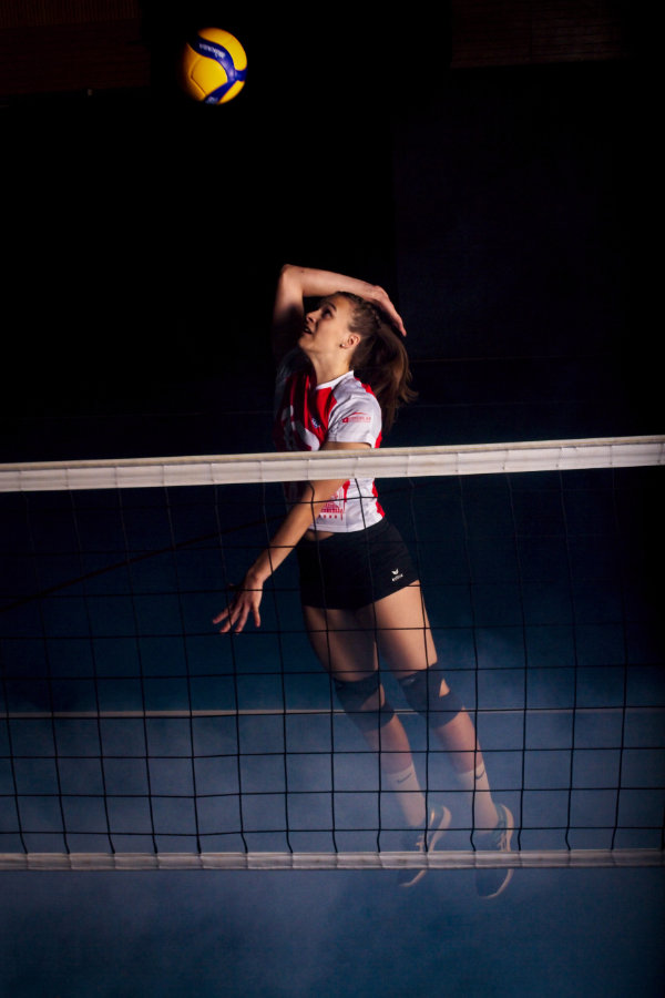 Volleyballerin mit angewinkeltem Angriffschlag hinter Netz, während am Boden Nebel zu sehen ist.