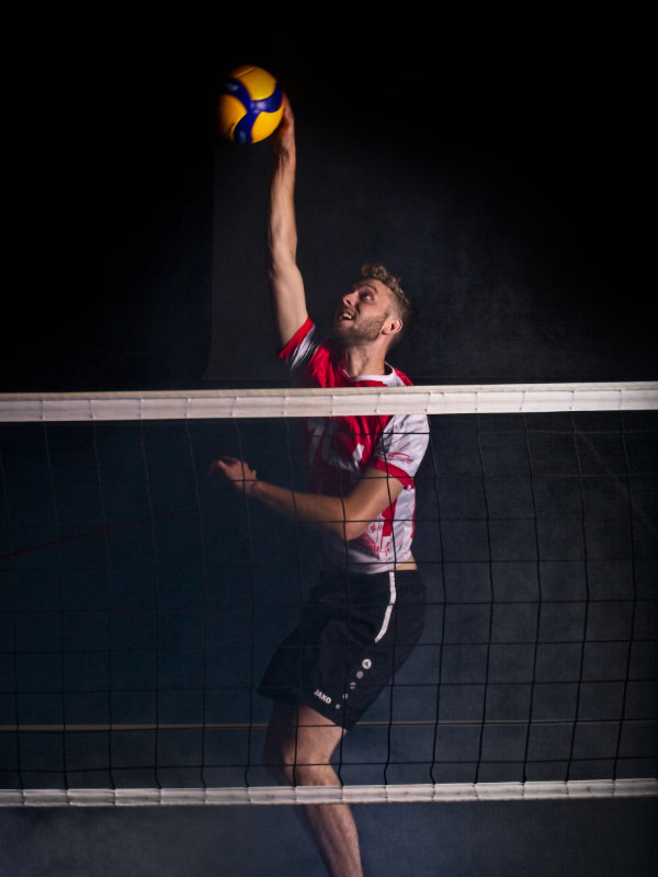Volleyballer mit ausgestrecktem Angriffschlag hinter Netz, während am Boden Nebel zu sehen ist.