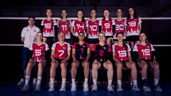 Klassisches Teamfoto in zwei Reihen eines Volleyball Team. Vordere Reihe sitzt auf einer Bank. Im Hintergrund ist ein Volleyballnetz zu sehen.