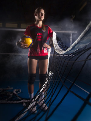 Frau posiert mit Volleyball vor einem Volleyballnetz. Über ihre Schultern liegt ein weiteres Volleyballnetz.